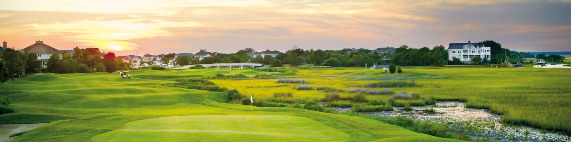 A scenic golf course with lush greens, surrounded by water and homes under a vibrant sunset sky.