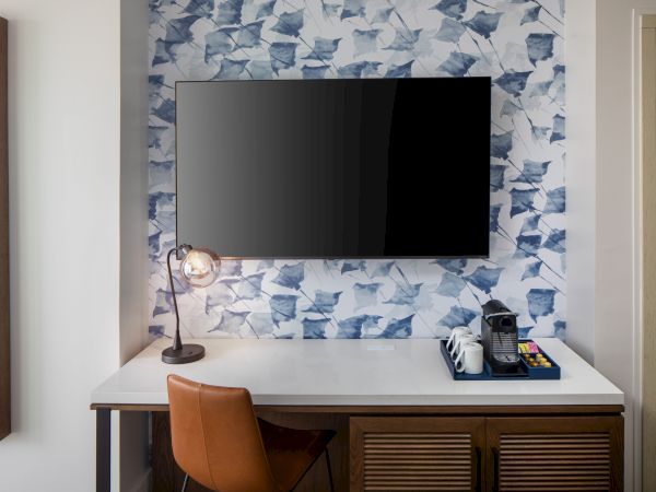 A modern setup with a TV on a patterned wall, desk, chair, lamp, and a coffee maker with supplies, creating a cozy workspace.