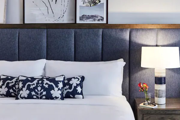 A bed with blue floral pillows sits against a padded headboard. A lit lamp and flowers are on a wooden nightstand, with art above.