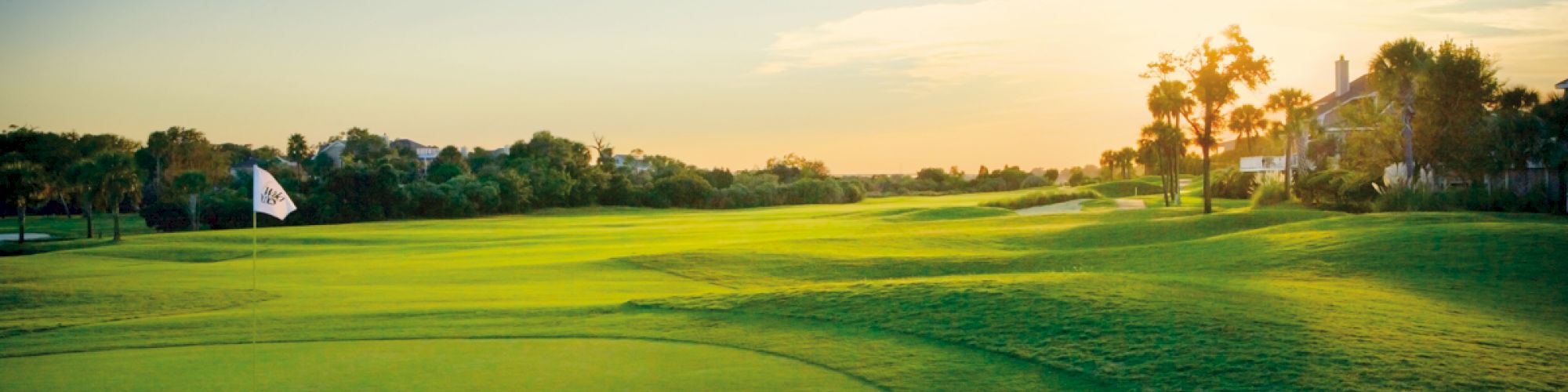 A scenic golf course at sunset with a flag indicating a hole, surrounded by trees and bushes, under a clear sky.