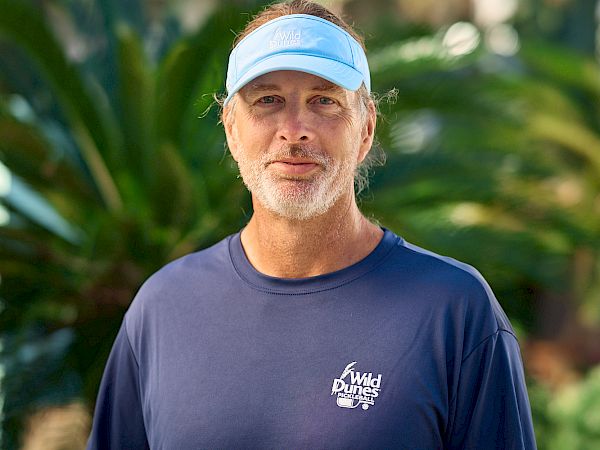 A man in a navy blue shirt and light visor stands outdoors, holding a black paddle ball racket, ready for a game.