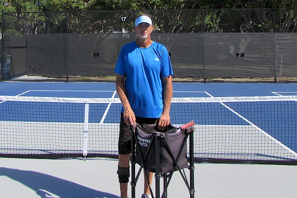 A person in sportswear stands on a blue pickleball court holding a chair; others play on nearby courts with trees in the background.