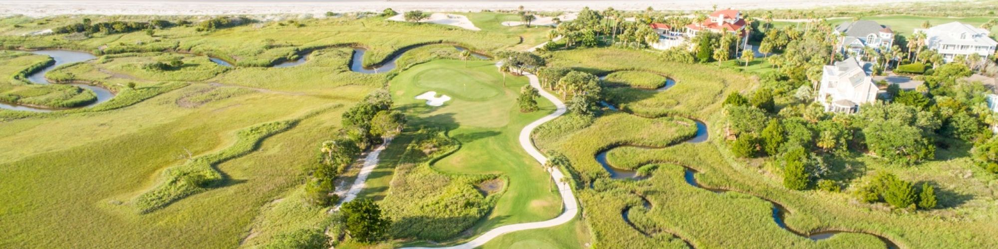 The image shows a coastal golf course with winding paths and water features, bordered by the ocean on one side and houses on the other.