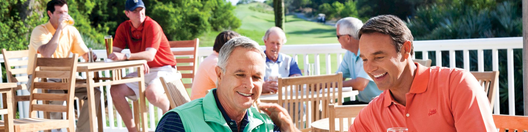 A group of people is enjoying a sunny day on a patio near a golf course, with drinks and smiles.