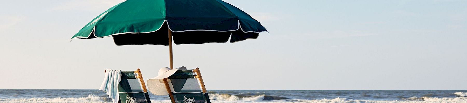 Beach scene with two wooden chairs under a green umbrella, a basket, waves in the background, and clear blue skies.