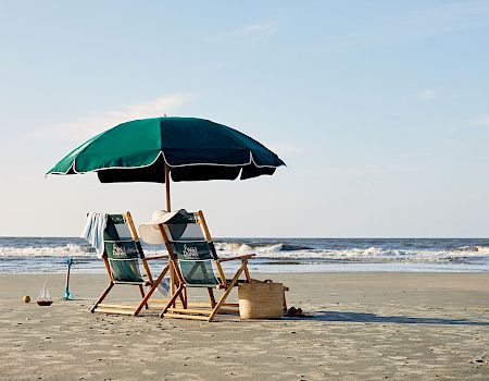 Beach scene with two wooden chairs under a green umbrella, a basket, waves in the background, and clear blue skies.