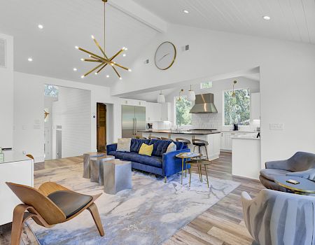 A modern living room with a blue sofa, stylish chairs, decorative lighting, and a view into a kitchen area, featuring sleek furnishings.