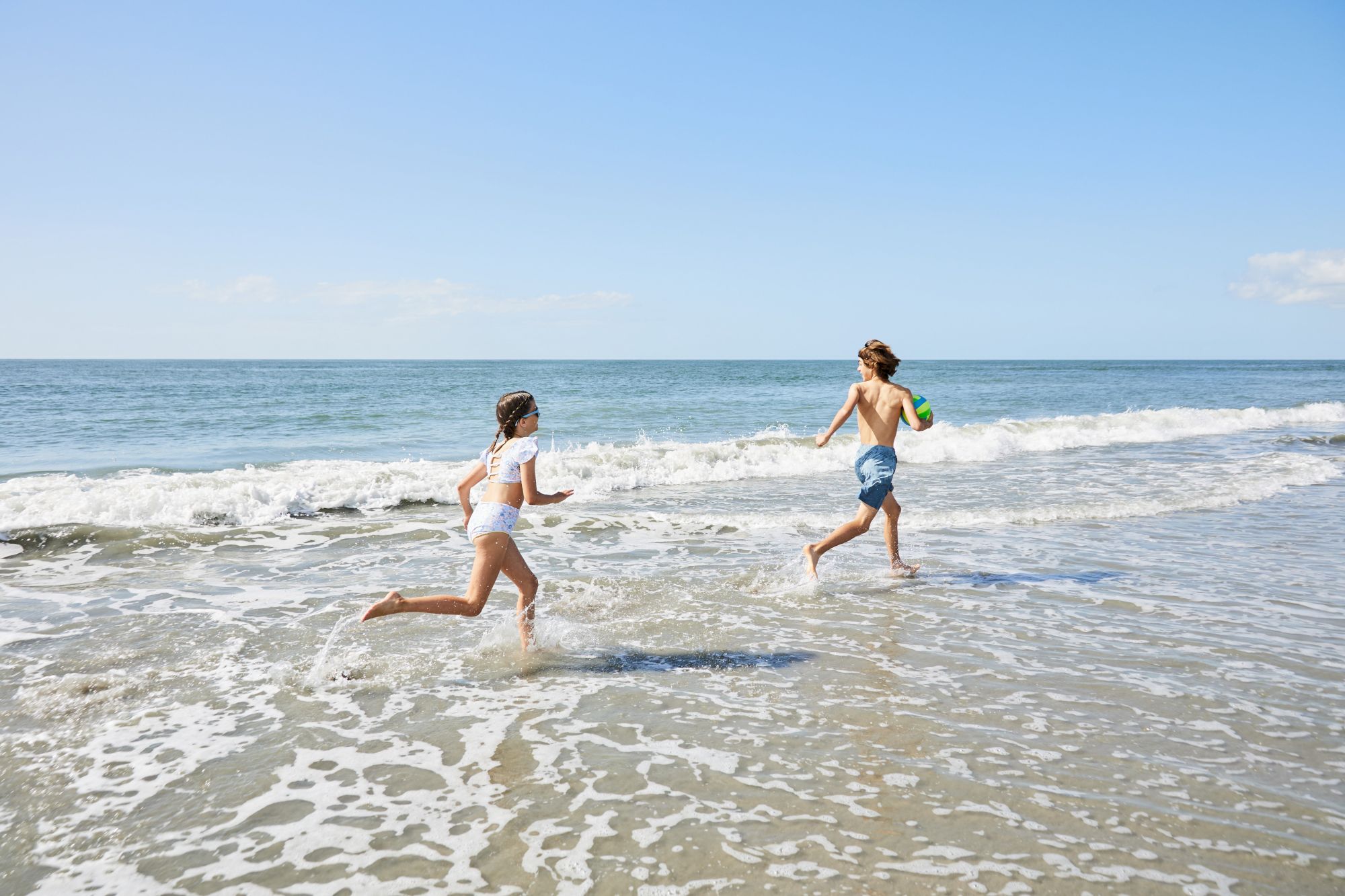 Two kids are playing and running along a beach shoreline with waves in the background on a clear, sunny day.