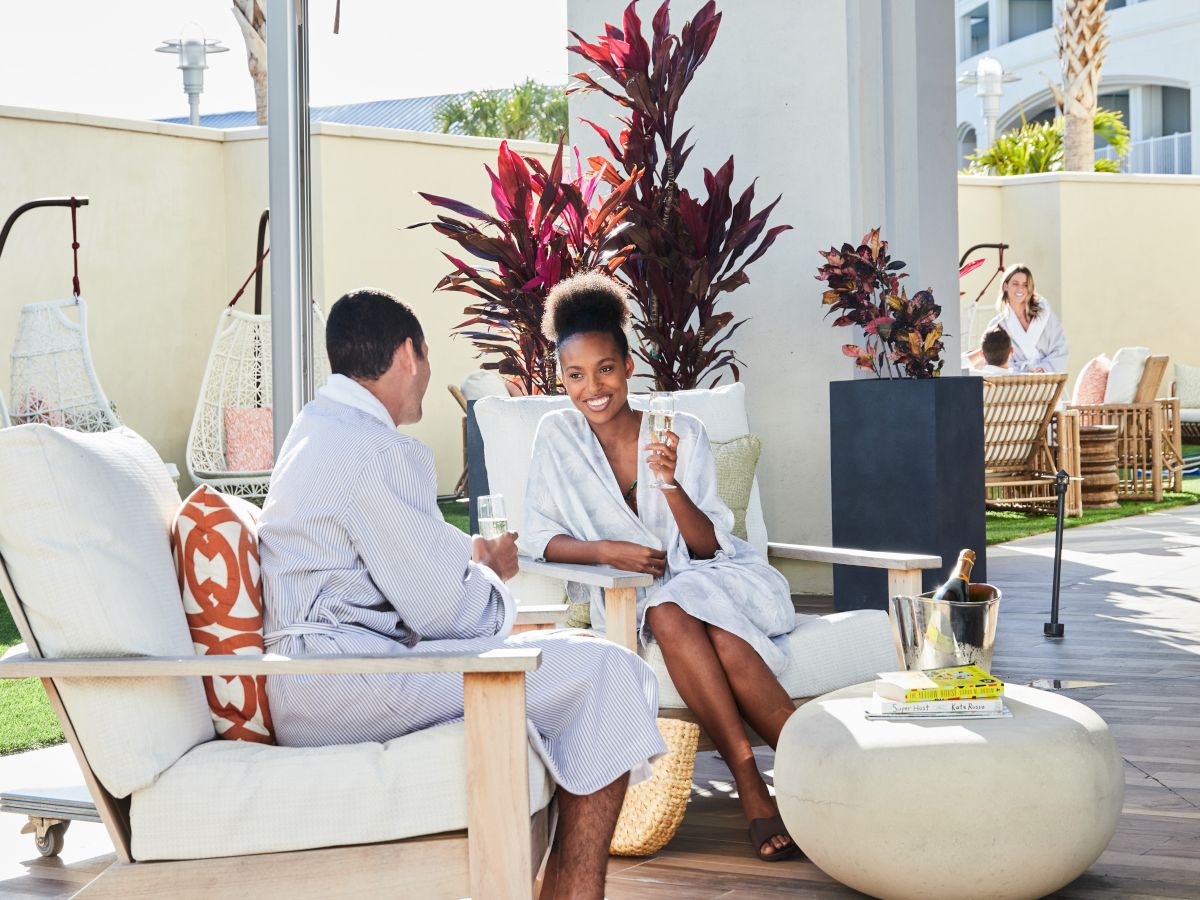 People relaxing outdoors in robes, seated on cushioned chairs, enjoying drinks; a serene setting with plants and swing chairs.
