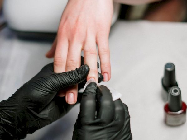 A person's nails are being cleaned or polished by someone wearing black gloves, with nail polish bottles nearby.