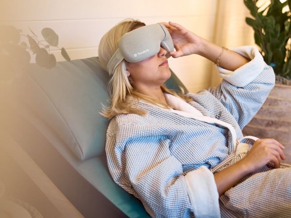 A woman lounges in a chair, wearing a fluffy robe and a white eye mask, relaxing with her hand resting on her lap and plants in the background.