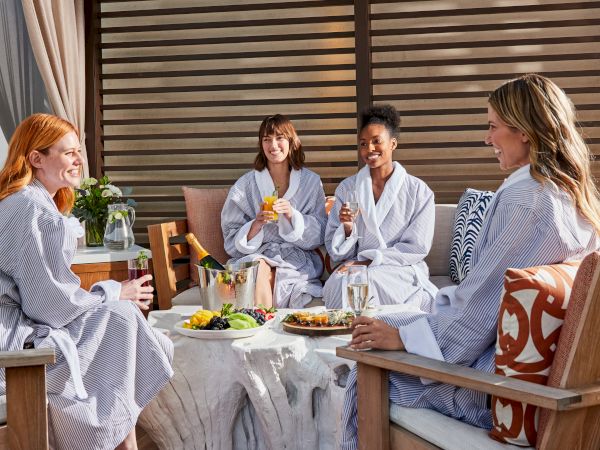 Four people in robes sit and enjoy drinks and snacks around a table, appearing relaxed and happy in a cozy setting.