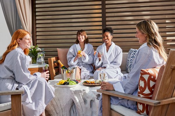 Four women in robes are enjoying drinks and snacks, seated around a table outdoors, with a relaxed and cheerful ambiance.