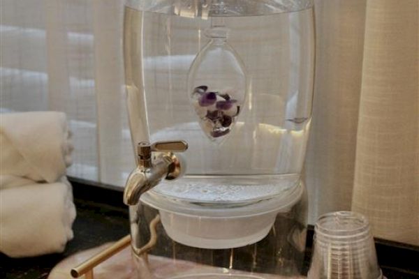 A water dispenser with a spout, infuser containing stones, plastic cups, and rolled towels, set on a table in front of sheer curtains.