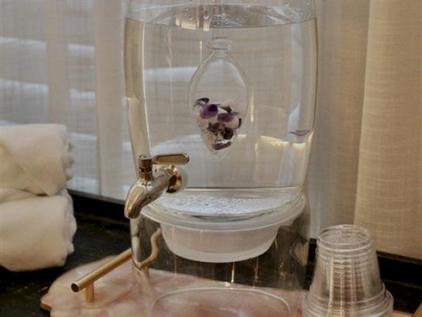 A water dispenser with a spout, infuser containing stones, plastic cups, and rolled towels, set on a table in front of sheer curtains.