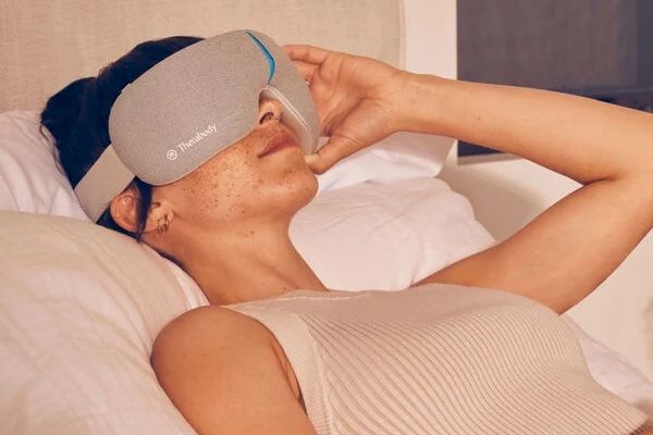 A person is lying on a bed wearing a gray eye massager device, appearing relaxed and resting comfortably.