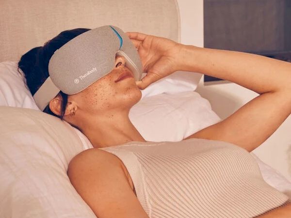 A person is lying on a bed wearing a gray eye massager device, appearing relaxed and resting comfortably.