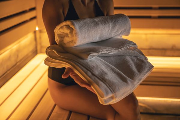 A person in a sauna holding folded towels, with warm lighting surrounding them.
