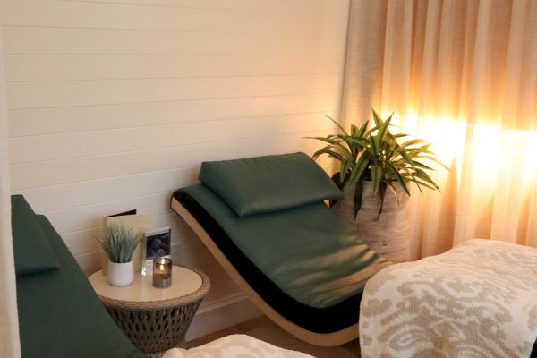 The image shows a spa room with two lounge chairs, blankets, a small table with candles, and a plant under soft lighting.