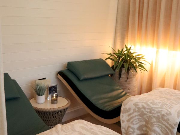 The image shows a spa room with two lounge chairs, blankets, a small table with candles, and a plant under soft lighting.