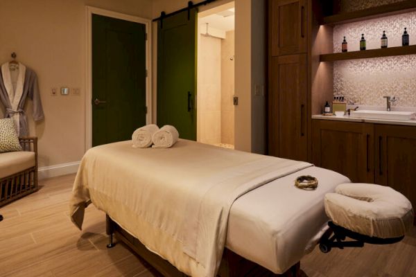 A tranquil spa room features a massage table, a robe on a chair, soft lighting, and a sink with bottles on shelves.