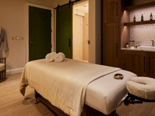 A tranquil spa room features a massage table, a robe on a chair, soft lighting, and a sink with bottles on shelves.