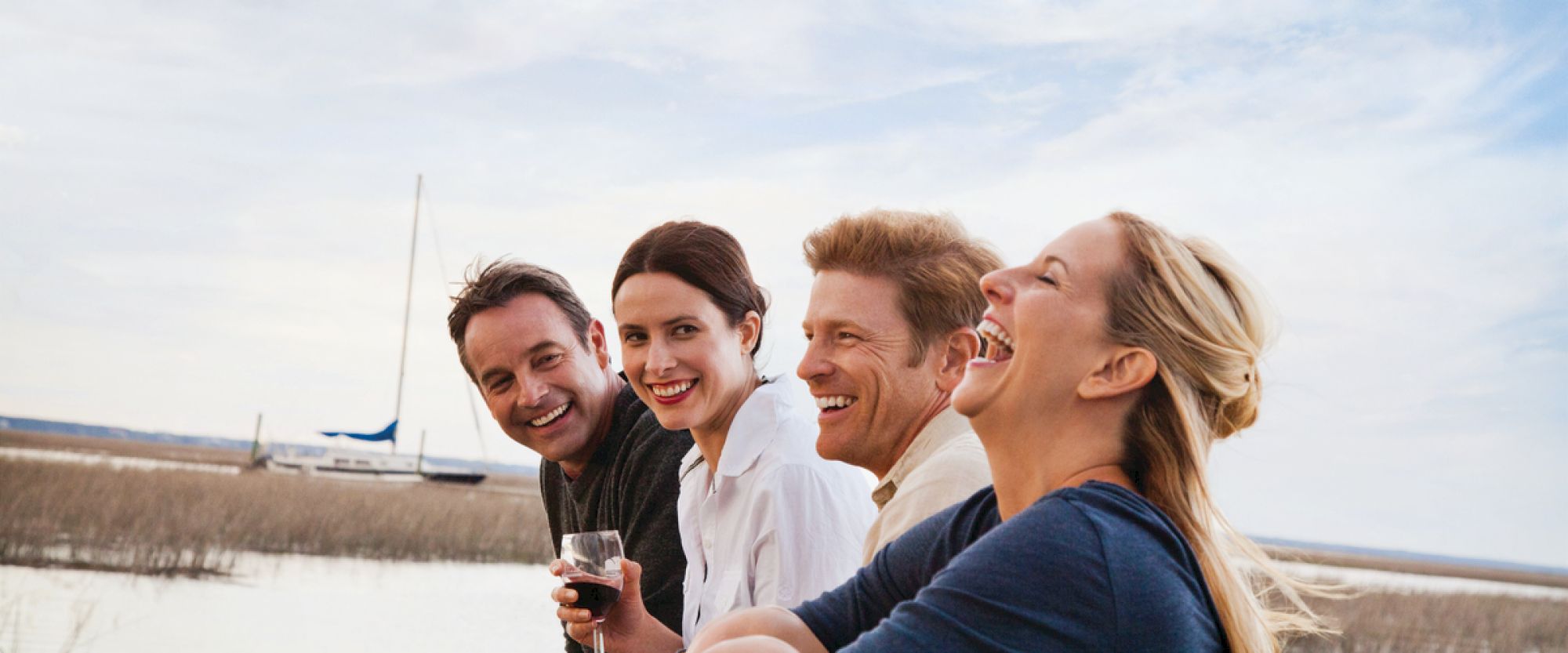Four people are laughing and enjoying themselves near a body of water with a sailboat in the background.