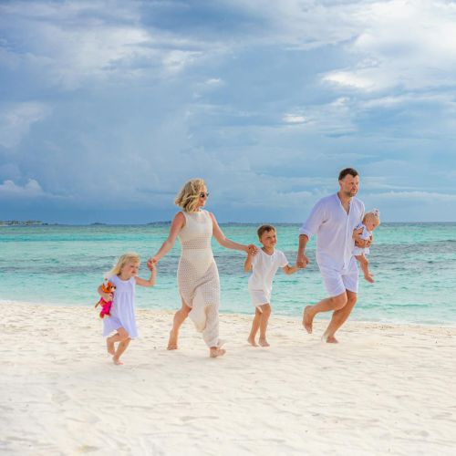 A family of five, including young children, walks hand-in-hand on a sandy beach by a turquoise sea under a partly cloudy sky.