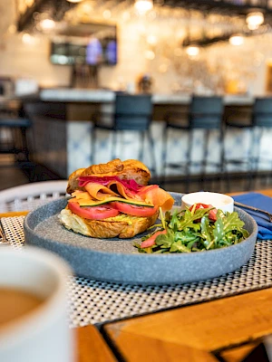 A sandwich with orange and pink layers, served with greens and sauce, sits on a plate in a cozy café setting.