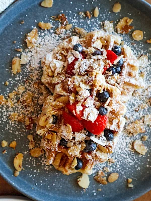 A plate of waffles topped with berries, powdered sugar, nuts, and syrup, presented on a dark plate.