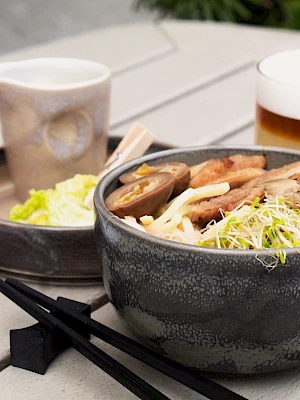 A bowl of ramen with sprouts, mushrooms, and egg, a drink, a sauce pitcher, a side dish, and utensils on a table.