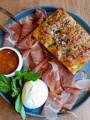 A plate with cured ham, burrata cheese, focaccia bread, a small dish of jam, and fresh basil leaves.