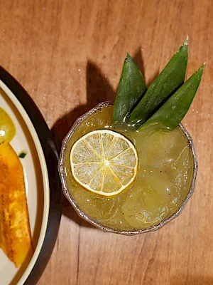 A dish with plantain chips and a green-colored cocktail garnished with lime and leaves on a wooden table.