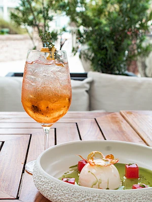 A refreshing orange cocktail and a dessert with sauce and cubes are on a wooden table outdoors.
