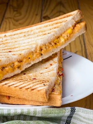 A grilled sandwich with cheese and corn, served with dips on the side, sits on a white plate over a wooden table.