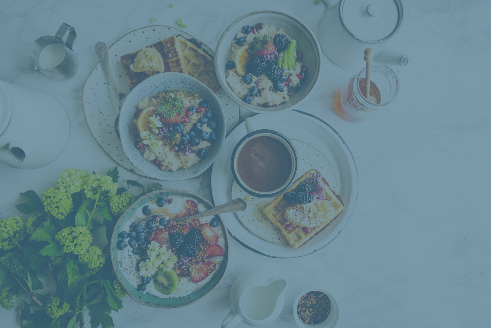 A breakfast spread with bowls of fruit and oatmeal, waffles, a pot of tea, milk, honey, and fresh flowers on the side.