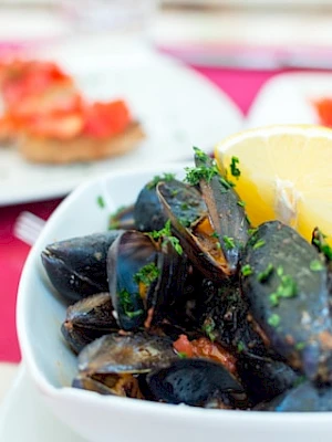 A bowl of mussels garnished with herbs and a lemon slice, with tomato bruschetta in the background on a red tablecloth.