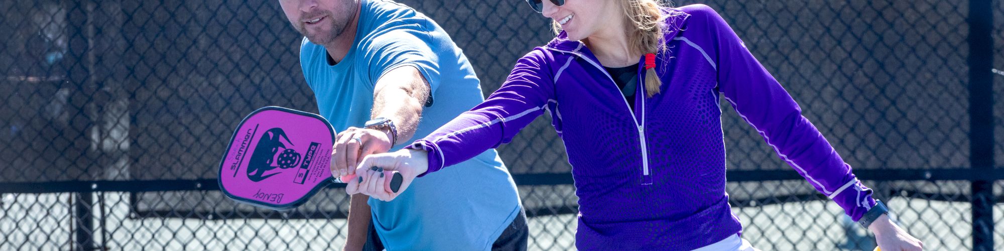 Two people are playing pickleball on a court. They are holding paddles and appear engaged in the game, wearing casual sports attire.
