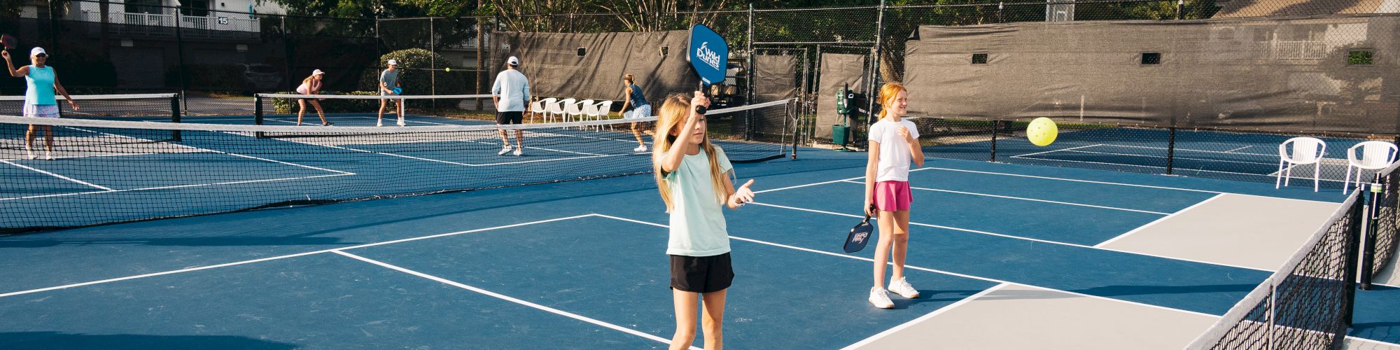 People are playing pickleball on blue courts with paddles and a yellow ball under clear skies with trees in the background.