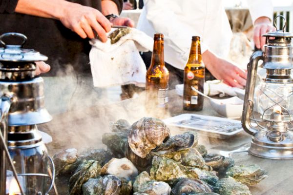 People are shucking oysters on a table with lanterns, beer bottles, and steam rising from the oysters.