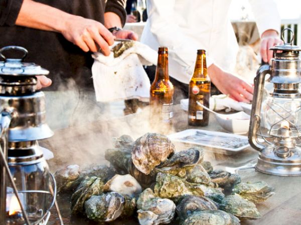 People are shucking oysters on a table with lanterns, beer bottles, and steam rising from the oysters.