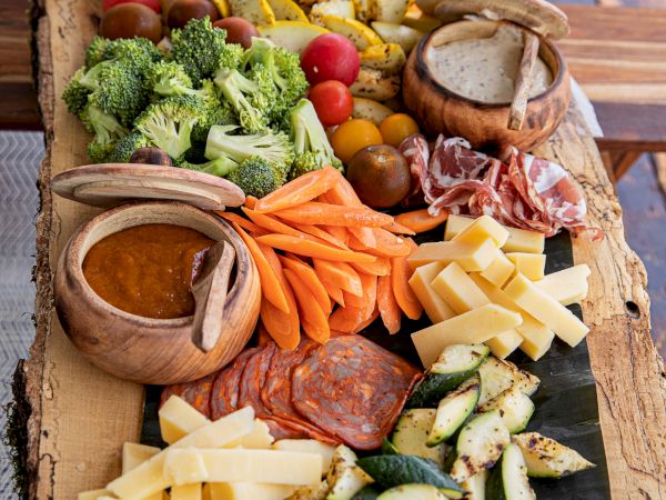 A wooden board with sliced vegetables, cheese, charcuterie, dips, and small bowls. Plates are stacked in the background on the table.