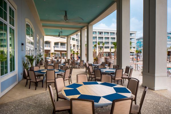 Outdoor restaurant seating area with round tables and chairs under a covered patio, overlooking a hotel pool and buildings.