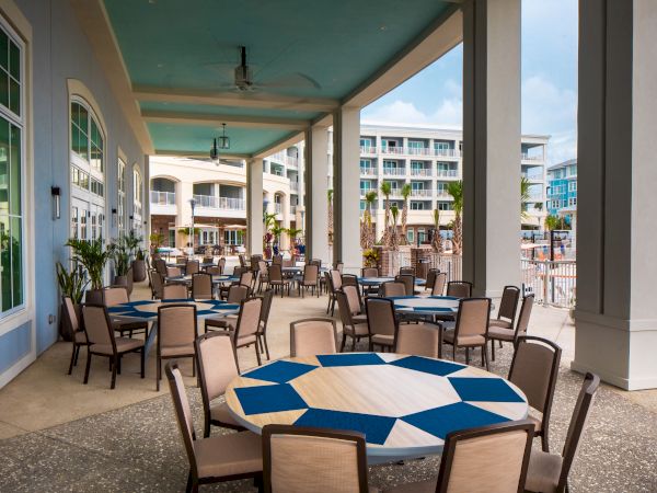 Outdoor restaurant seating area with round tables and chairs under a covered patio, overlooking a hotel pool and buildings.