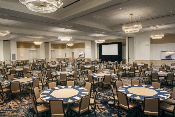 A large, empty banquet hall with round tables and chairs, chandeliers, and a projection screen at the front of the room.