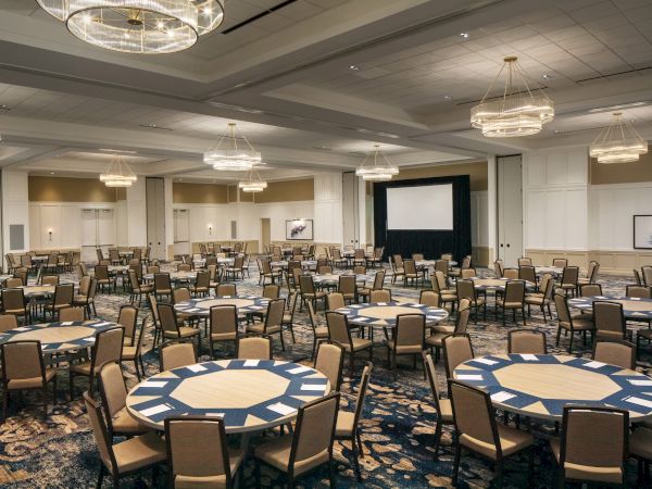 A large, empty banquet hall with round tables and chairs, chandeliers, and a projection screen at the front of the room.