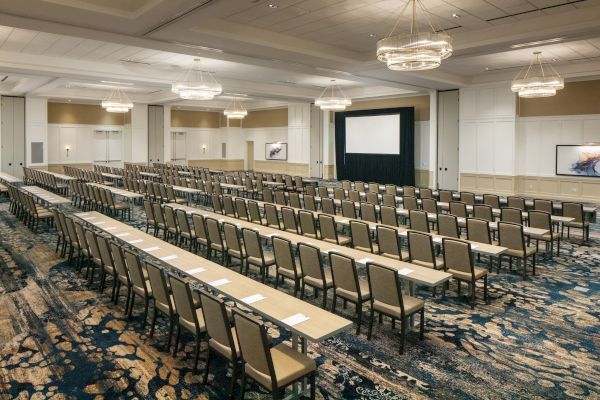 The image shows a conference room with rows of desks and chairs facing a large screen at the front.