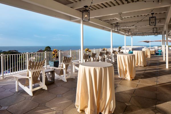 An outdoor patio with covered tables, Adirondack chairs, string lights, and ocean views.