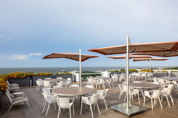 A rooftop patio with round tables and white chairs under large red umbrellas overlooking the ocean and lined with orange flowers.