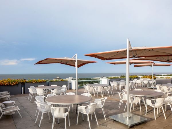 A rooftop patio with round tables and white chairs under large red umbrellas overlooking the ocean and lined with orange flowers.